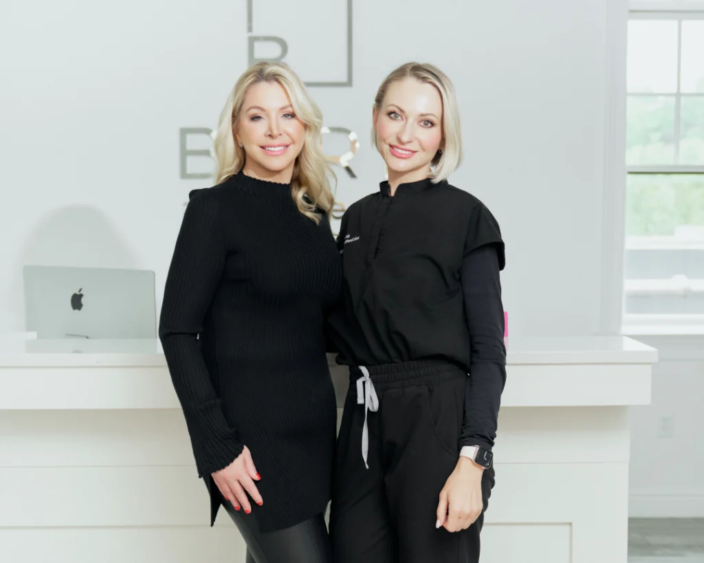 Two smiling women at a medical weight loss clinic, one in black attire and the other in a medical uniform, standing at the reception desk.